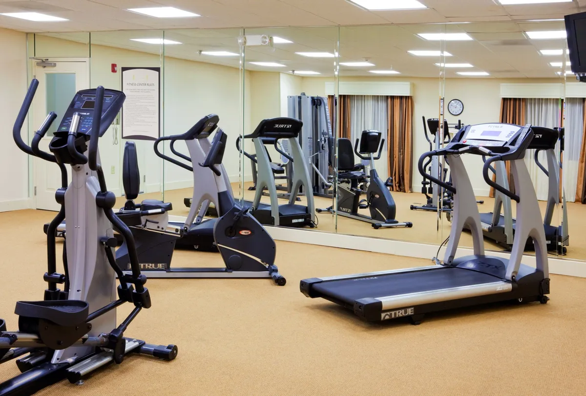 A modern, well-lit fitness center is depicted, showcasing an elliptical machine, exercise bike, treadmill, and mirrored walls. The scene is active and motivational, inviting guests to maintain their wellness routines in a fully equipped space available at any time.