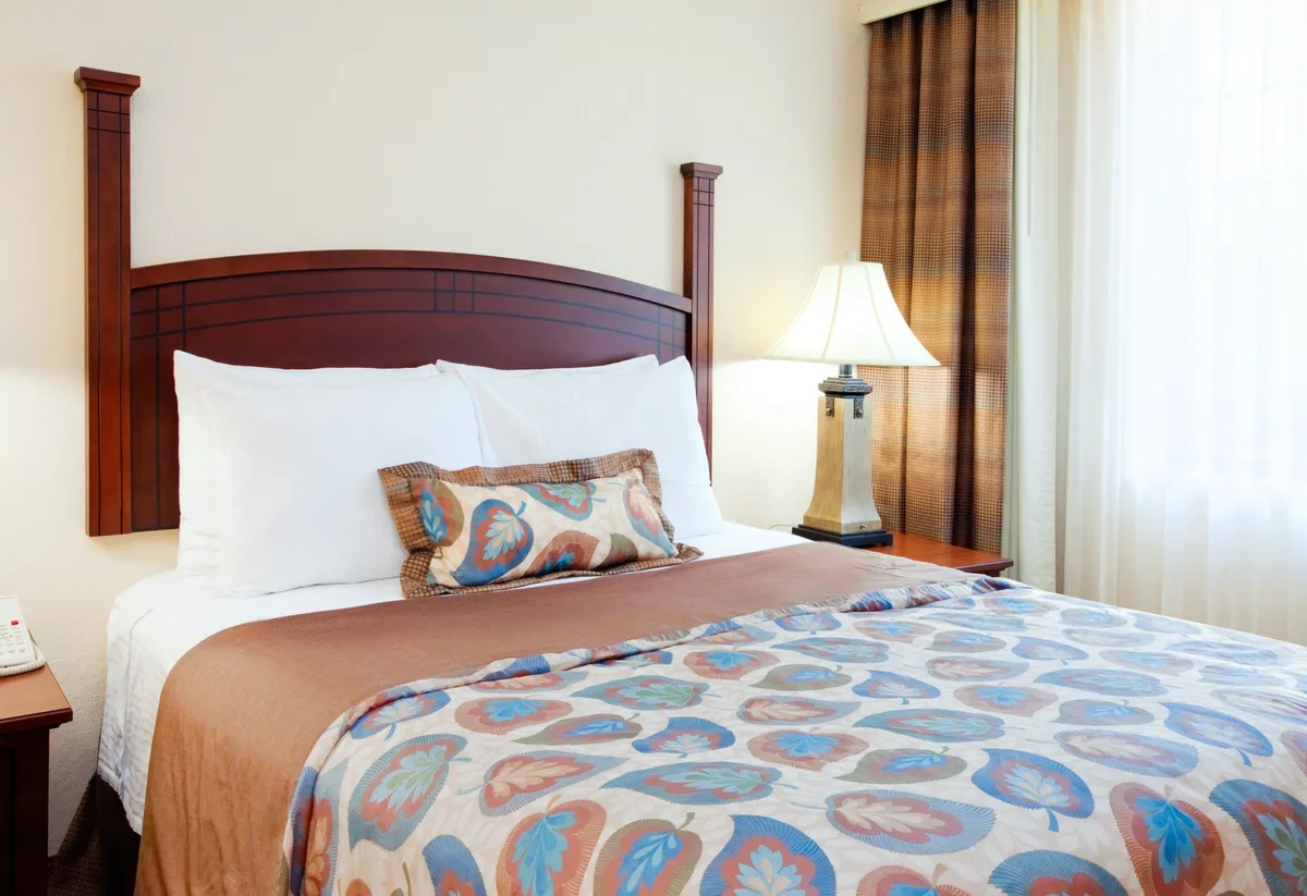 A serene bedroom with a comfortable bed, decorative pillows, a lamp, and soft curtains softly filtering in daylight. The atmosphere is tranquil and designed for restful sleep or peaceful reading.