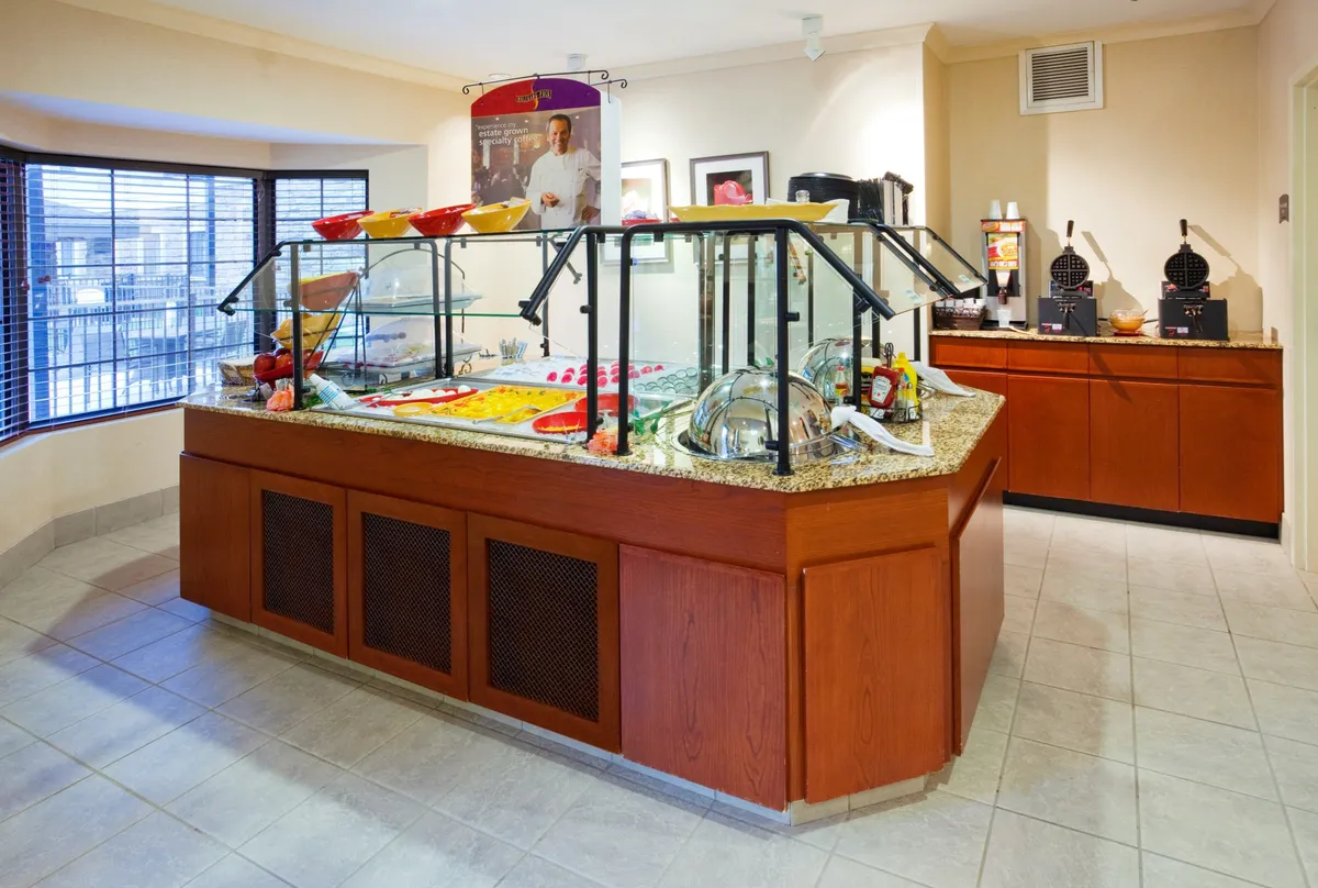 A bright, inviting breakfast area featuring a food display counter with colorful bowls and glass serving containers. The space includes coffee and waffle stations, encouraging leisurely meals and social interaction. Guests are seen enjoying breakfast and mingling in the comfortable, communal setting.