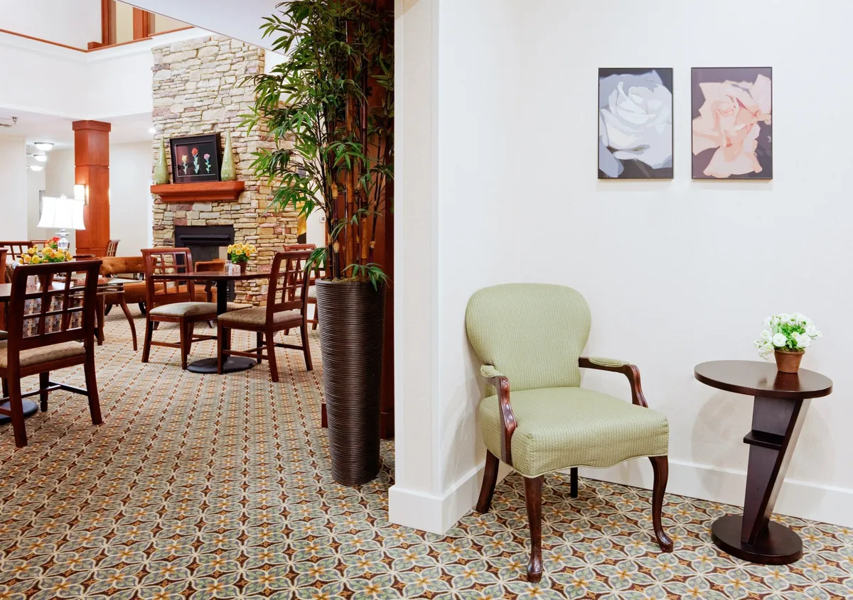 A comforting and stylish lounge area featuring a plush green chair and small round table, accented by modern artwork, a large potted plant, and a fireplace in the background. The space feels warm and inviting, ideal for quiet relaxation or intimate conversations.