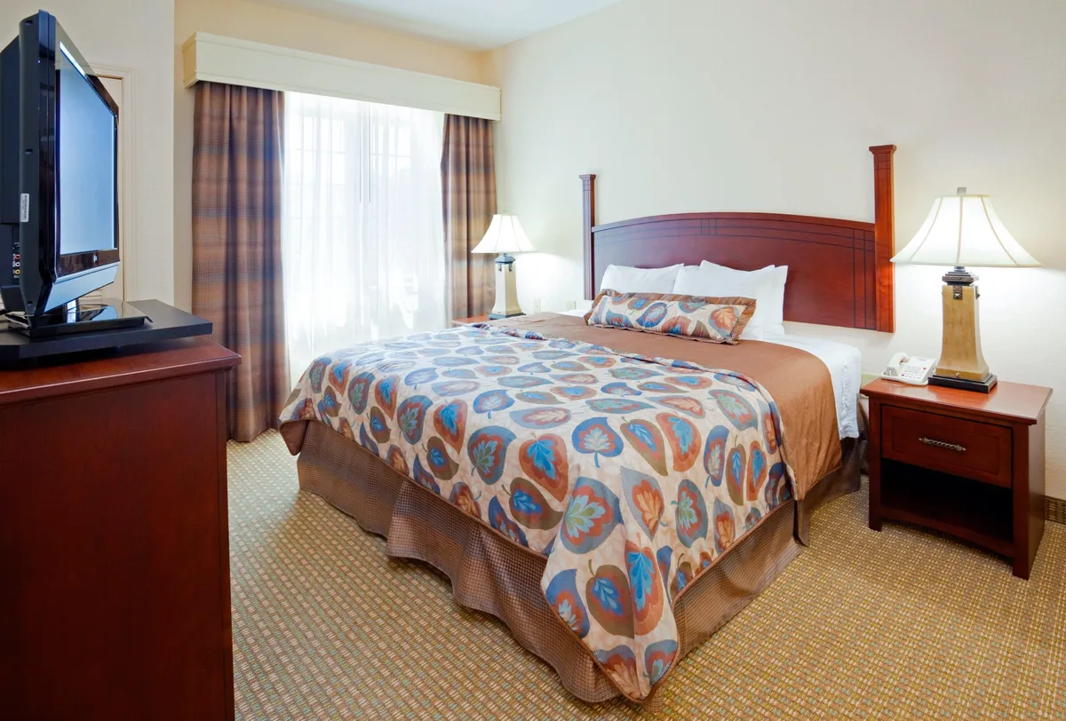A cozy bedroom featuring a bed with patterned bedding, soft lamps on either side, a television across from the bed, and a window with drawn curtains letting in gentle natural light. The space feels inviting, perfect for relaxing, reading, or enjoying a quiet morning.