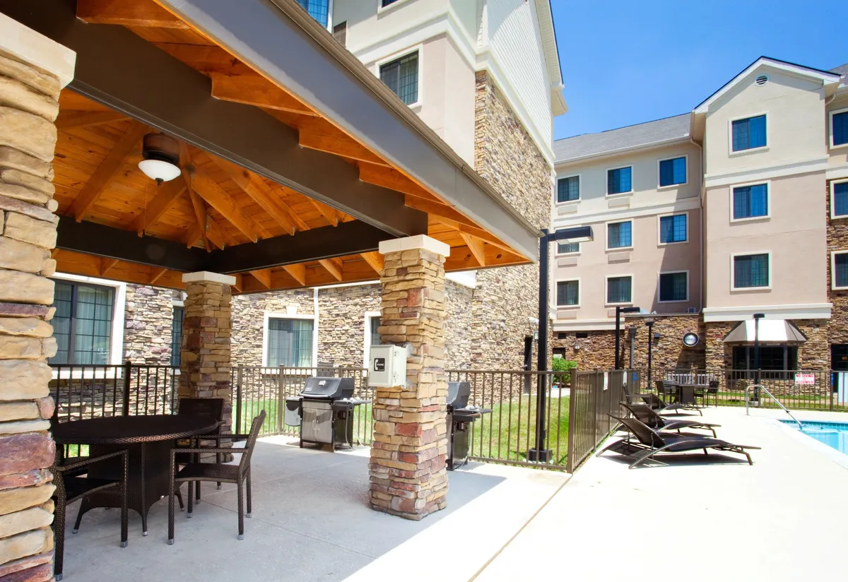 An exterior BBQ and outdoor seating area featuring stone pillars, grill stations, and a view of the swimming pool. The space is set up for outdoor dining, socializing, and enjoying a classic cookout with friends or family.