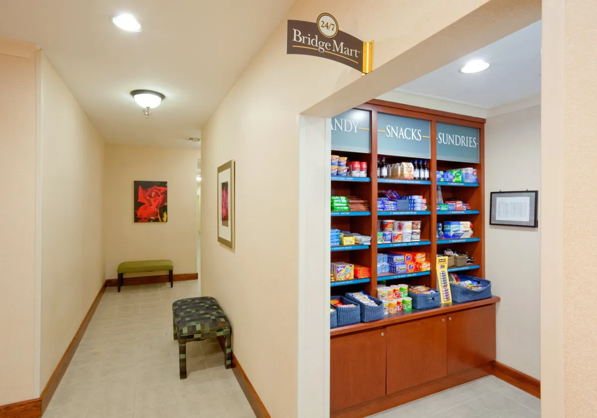 A cozy interior scene featuring the Bridge Mart snack shelf, a welcoming seating area, and casual guests either selecting snacks or relaxing. The ambiance is casual and inviting, with artwork on the wall contributing to the comfortable atmosphere. The space feels like a convenient common area for guests starting their day or grabbing a quick bite.