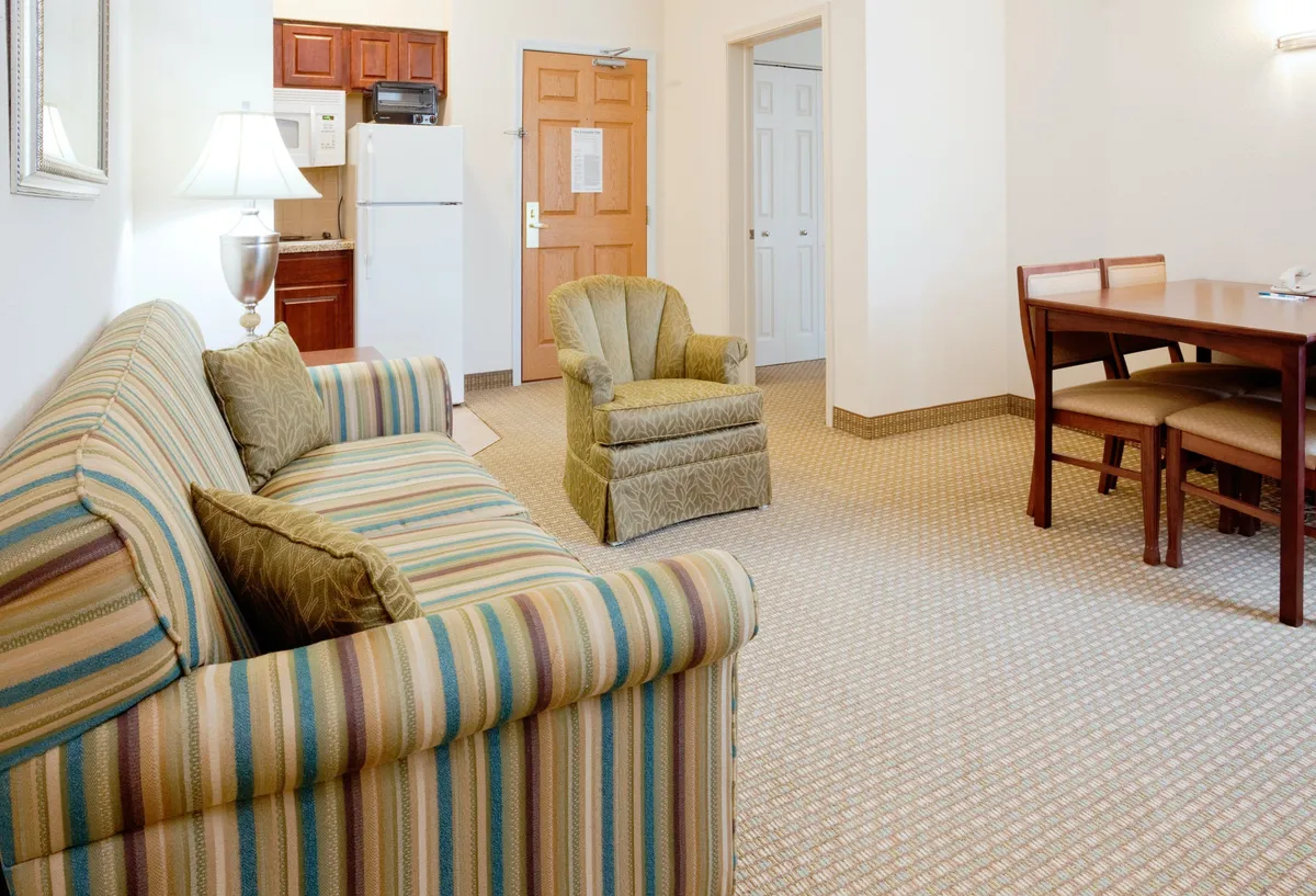The image shows a welcoming, open-plan condo living space featuring a plush striped sofa, a vibrant green chair, and a dining table set for enjoying meals together. The inviting furnishings and soft lighting create a warm, relaxing atmosphere ideal for socializing and unwinding after exploring Durham or visiting nearby universities.