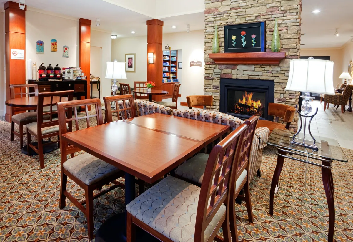 A cozy, open-plan lounge area with a fireplace, wooden tables, comfortable seating, and local artwork on the walls. The room is arranged to encourage conversation and relaxation, perfect for gatherings over coffee or casual meals.