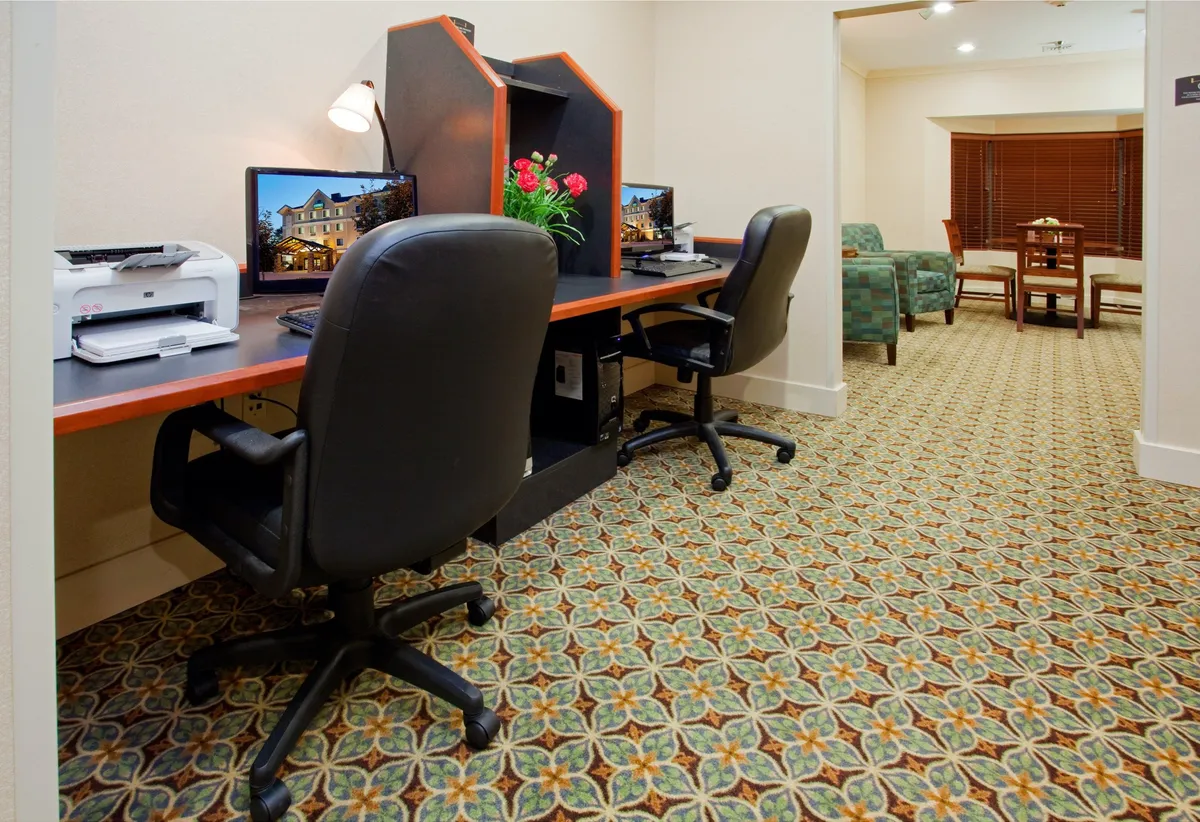 This image features a professional-yet-inviting business center setup, complete with desks equipped with computers, a printer for guest use, and a floral arrangement that adds a welcoming touch. The space is organized and conducive to productivity, offering guests a dedicated area for work and business-related needs.